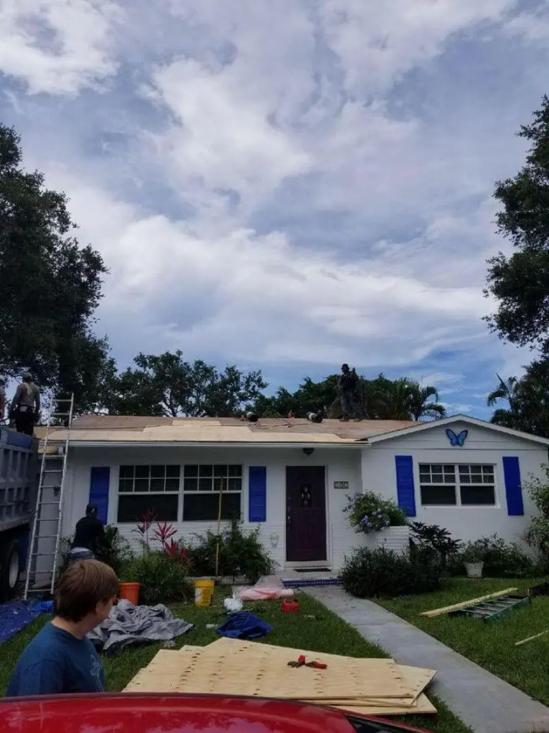 Small white cottage with blue shutters mid re-roof, new plywood decking stacked on the lawn