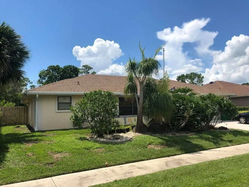 Ranch home with a brown shingle roof behind palms under a blue sky